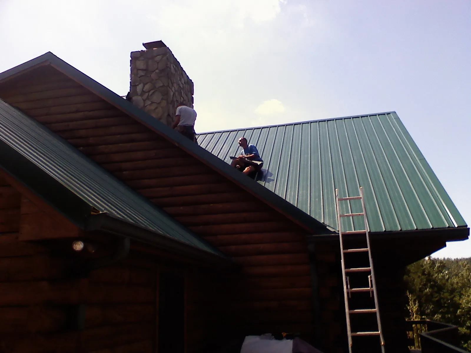 Roofers installing green metal panels beside a stone chimney on a log home in Cedarville